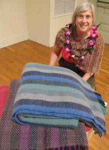 shows weaver wearing lei kneeling behind pile of blankets