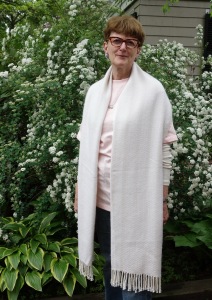 Susan wearing white shawl draped around her shoulders and hanging down straight in front. Backdrop of green shrubs with white flowers.