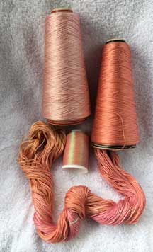 Two cones of yarn at the top in pink and light coral, with a small spool of coral iridescent thread in the middle and partial skein of pink and coral hand-dyed silk yarn at the bottom.